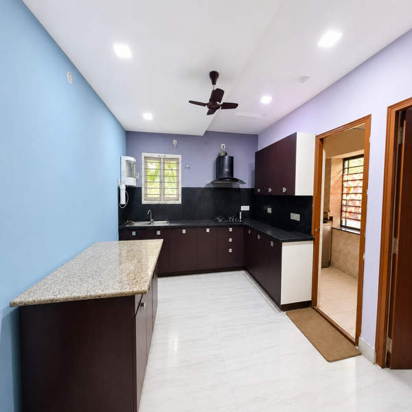 L-shaped kitchen, modular kitchen, modern kitchen, contemporary kitchen, spacious kitchen, home kitchen, residential kitchen, rental home kitchen, kitchen interior, kitchen cabinetry, dark wood cabinets, granite countertop, kitchen island, breakfast counter, kitchen peninsula, black backsplash, marble flooring, kitchen sink, stainless steel sink, kitchen faucet, chimney hood, cooking area, built in storage, overhead cabinets, lower cabinets, kitchen drawers, kitchen workspace, organized kitchen, functional kitchen, well lit kitchen, ceiling lights, ceiling fan, blue accent wall, lavender wall, elegant kitchen, stylish home interior, practical kitchen design, family kitchen, bachelor accommodation, student accommodation, students, business professionals, working professionals, office goers, corporate housing, rental accommodation, modern living space, urban home, independent kitchen, fully equipped kitchen, comfortable home, move in ready property, residential interior.